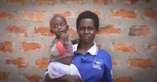 Äiti seisoo pieni lapsi kädessään tiiliseinän edessä.. Uganda.