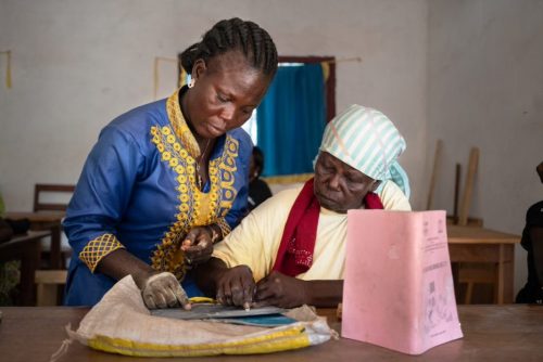 Afrikkalainen naisopettaja ohjaa vanhempaa naista tekstintuottamisessa liitutaululle.