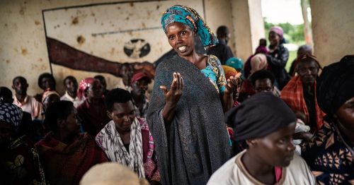 Afrikkalainen nainen seisoo keskellä istuvia naisia ja selittää asiaansa katsoen kameraan.