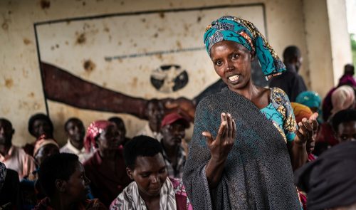 Afrikkalainen nainen seisoo keskellä istuvia naisia ja selittää asiaansa katsoen kameraan.