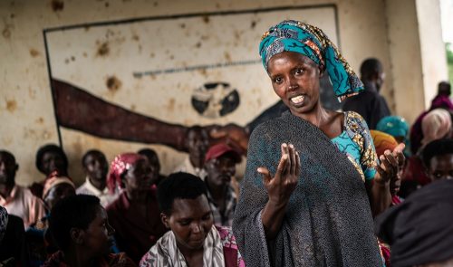 Afrikkalainen nainen seisoo keskellä istuvia naisia ja selittää asiaansa katsoen kameraan.