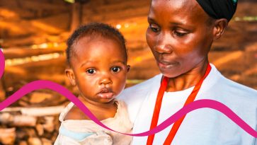 Ugandalainen nainen valkoisessa t-paidassa seisoo pieni vauva sylissään. Vauva katsoo suoraan kameraan ja äiti katsoo lasta pieni hymy kasvoillaan.