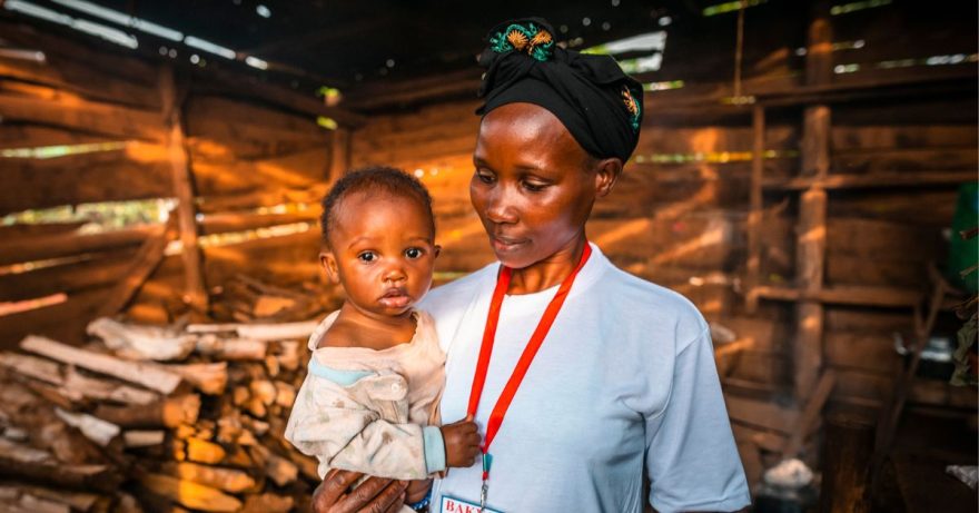 Ugandalainen nainen valkoisessa t-paidassa seisoo pieni vauva sylissään. Vauva katsoo suoraan kameraan ja äiti katsoo lasta pieni hymy kasvoillaan.