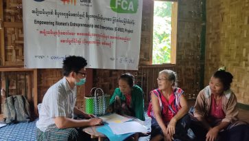A group of four people sit on the floor of a room with bamboo walls around a small table covered in papers. They are talking. On the wall hangs a banner with writing in Burmese and the logos of the Kaw Lah Foundation, Women's Bank and FCA