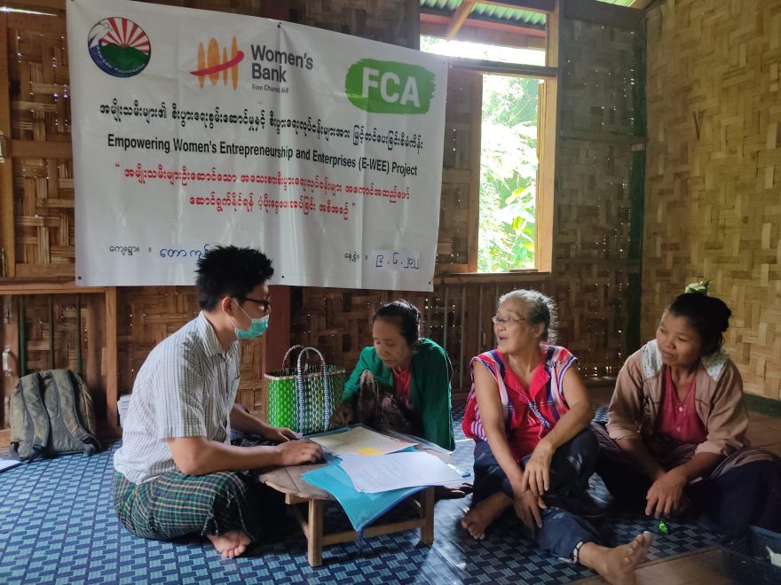 A group of four people sit on the floor of a room with bamboo walls around a small table covered in papers. They are talking. On the wall hangs a banner with writing in Burmese and the logos of the Kaw Lah Foundation, Women's Bank and FCA