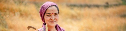 Nepalilailen nainen hymyilee ja katsoo suoraan kameraan. Hän seisoo viljapellolla sirppi kädessään.