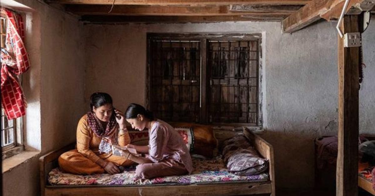 Nepalilaiset nainen ja tyttö istuvat sängyllä. Tyttö lukee ja äiti kuuntelee vieressä.