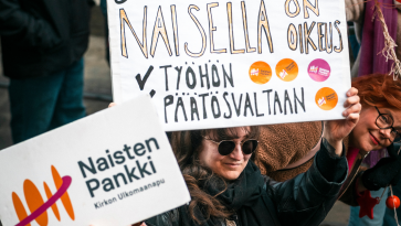 nainen pitää päänsä yllä kylttiä, jossa lukee naisella on oikeus työhön ja päätösvaltaan ja kyltin takaa kurkkaa toinen nainen.
