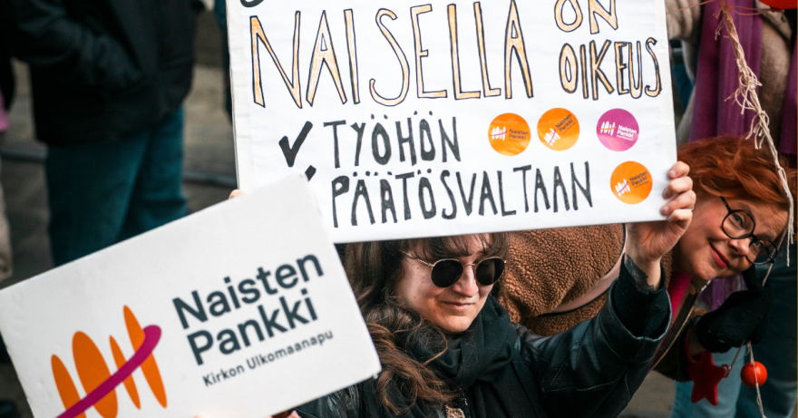 nainen pitää päänsä yllä kylttiä, jossa lukee naisella on oikeus työhön ja päätösvaltaan ja kyltin takaa kurkkaa toinen nainen.